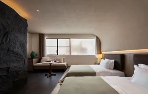 Modern hotel bedroom interior with two beds dressed in white and sage green, a large natural rock wall on the left, textured plaster ceiling, built-in daybed by the window, and warm ambient lighting.