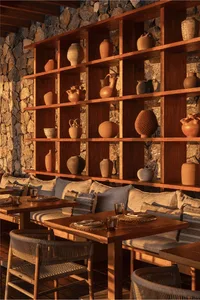 Cozy rustic dining area featuring a built-in wooden shelving unit filled with terracotta pottery, set against a natural stone wall with cushioned banquette seating and woven chairs.