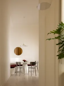 Modern minimalist dining area featuring a curved white banquette with red velvet upholstery, a white pedestal table, transparent acrylic and stainless steel chairs, a white pendant lamp, and a round brass mirror on the wall.