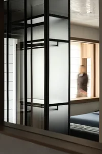 View through a black steel framed sliding partition with frosted glass panels into a modern minimalist bedroom with blue bedding and wooden window seat.