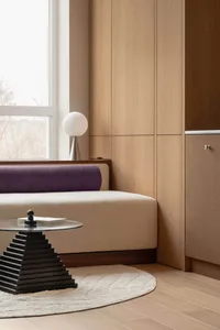 Modern living room corner featuring a purple upholstered window seat, light oak built-in storage, spherical table lamp, and a black geometric coffee table on a cream rug.