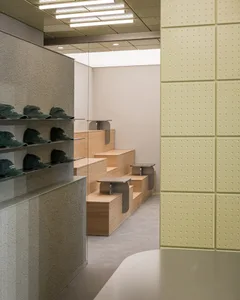 Modern retail interior with pale yellow perforated acoustic wall panels, concrete-textured shelving displaying green baseball caps, and light oak tiered steps with metal display tables.