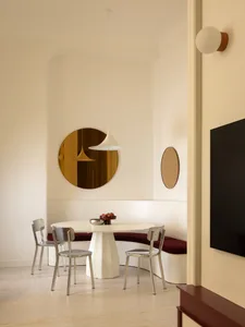 Modern dining area featuring a curved white banquette with deep red upholstery, a central white pedestal table, brushed steel chairs, a large round amber mirror, and a white pendant lamp.