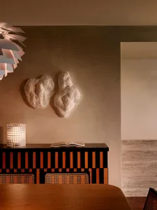 Warm minimalist dining area featuring a black and walnut striped console table, two textured organic wall sculptures, a white grid lantern, and a pleated pendant lamp.