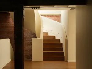 View of a modern curved wooden staircase with white plaster walls and an exposed red brick wall in the background, framed by a dark wooden beam in the foreground.