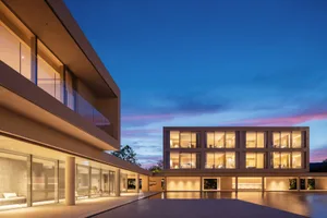 Modern two-story concrete house with extensive glass windows glowing warmly at twilight, featuring a large reflecting pool in the foreground and a clear blue-to-pink gradient sky.