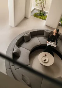 High-angle view of a modern living space featuring a large curved grey boucle sectional sofa surrounding a round coffee table, with a woman reading a book, opening to a lush sunken courtyard planted with moss, rocks and a Japanese maple tree.