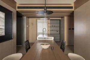 Modern minimalist dining room featuring a long oak wood table, black lattice-back chairs, textured boucle stools, and a dramatic black pendant light hanging above the table, with integrated kitchen cabinetry and wood slat ceiling in the background.