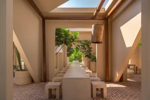 Long concrete dining table with matching stools arranged in a sunken courtyard under a skylight, surrounded by potted fiddle-leaf fig trees and warm beige walls with wooden beams.
