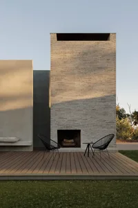 Modern outdoor deck featuring a tall light-colored brick fireplace, two black Acapulco chairs, a small side table, and a built-in concrete bench with cushion, surrounded by grass and trees.
