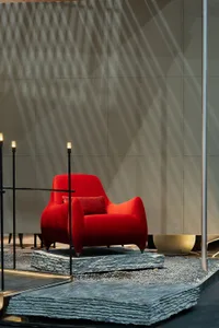 A bright red velvet armchair with a matching cushion sits on a raised platform made of stacked grey slate slabs, set against a large concrete wall with strong diagonal shadows and three tall black floor lamps nearby.