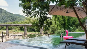Infinity swimming pool beside a river with two women swimming, pink flamingo float, lounge chairs under a large umbrella, surrounded by dense green trees and mountains in the background.