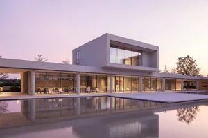 Modern minimalist concrete villa with large glass windows and an expansive reflecting pool at twilight, showing perfect symmetry between architecture and water.