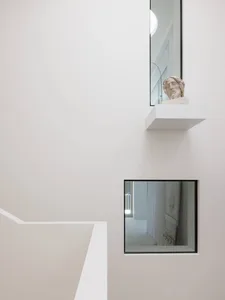 Minimalist white interior featuring a floating concrete shelf with a classical stone bust, viewed from a staircase with square black-framed window below.