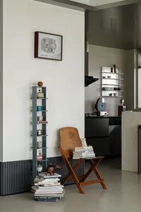 Modern minimalist living room corner featuring a wooden folding chair used as a side table, a tall blue open shelving unit, stacks of books, and a framed architectural floor plan on the wall.