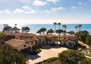 Aerial view of a luxurious Mediterranean-style stone villa with terracotta tile roofs, circular driveway, and lush landscaping overlooking the blue Pacific Ocean under a clear sky.