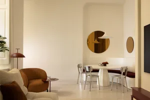 Modern dining area with a curved white pedestal table, built-in burgundy banquette, chrome chairs, a large round brass mirror, and a burnt orange boucle armchair in the foreground.