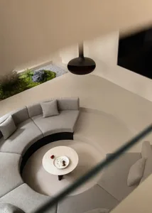 High-angle view of a modern living room featuring a large curved grey boucle sofa arranged around a round marble coffee table, with an indoor garden of ferns and rocks in the background and a black pendant lamp hanging above.