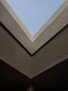 Looking straight up at a modern V-shaped concrete ceiling with a large rectangular skylight opening to blue sky.