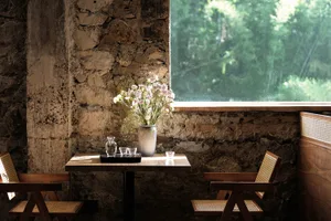 A rustic cafe dining nook featuring a rough exposed stone wall, a wooden table with fresh flowers, two wooden armchairs with rattan seats, and a large window overlooking green trees.