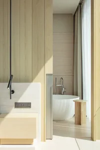 View into a serene minimalist bathroom with light oak wood paneling, a white freestanding bathtub, wooden side table, and sheer curtains allowing natural light.