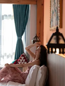 一位黑髮女子穿著粉色洋裝斜靠在鋪有白色枕頭的床上，一手枕在腦後，閉眼微笑，背景是溫馨的木質牆面、青綠色窗簾與暖光燈籠。