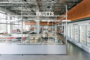 Wide view of a modern industrial-style grocery store featuring a large stainless steel central display island, glass refrigerated cases, concrete floors, and exposed ceiling infrastructure with a prominent FRUITS sign.