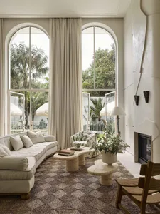 Bright living room featuring a curved white sofa, patterned rug, organic travertine coffee tables, a tropical print armchair, and large arched windows overlooking a palm-filled garden.