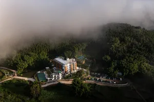 航拍照片顯示一座結合混凝土與玻璃立面的多層現代度假村，坐落於茂密森林與濃厚白霧的山區，周圍可見游泳池與停車場。