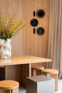 Modern dining area featuring a light birch plywood wall with mounted black speakers, a matching birch table, cylindrical wooden stools, a metallic cube stool, and a large silver foil vase filled with yellow forsythia branches.