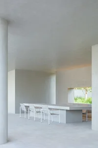 Minimalist concrete dining room featuring a long built-in concrete table, white metal-framed chairs, and a large horizontal window overlooking a green garden.