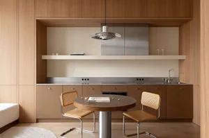 Modern minimalist kitchen and round dining table with two cane chairs, warm oak cabinetry, stainless steel countertop and range hood, floating shelf, and sculptural pendant light.