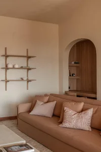 Modern minimalist living room with a terracotta fabric sofa, patterned cushions, floating wooden shelves, and a built-in arched wooden niche desk area.