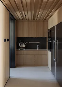 Modern narrow butler pantry with light oak wood cabinetry, black stone accent wall, wood slat ceiling, built-in coffee station, and stainless steel refrigerator.