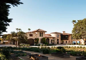 Expansive Tuscan-style stone villa with terracotta roof, arched windows, lush Mediterranean garden and distant ocean view under clear blue sky.
