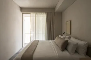 Minimalist bedroom interior with a large bed dressed in white and beige linen bedding, a woven throw, sheer and heavy curtains framing a glass door, and a simple framed artwork on the wall.