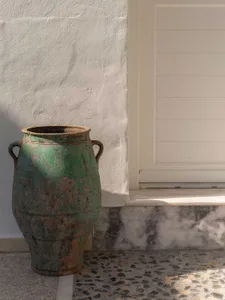 Large antique green ceramic urn with two handles placed on a terrazzo floor next to a white textured wall and marble base.
