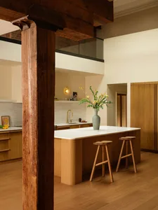 Modern kitchen featuring a curved oak island with white marble countertop, two wooden stools, brass faucet, and a large exposed dark wood beam in the foreground.