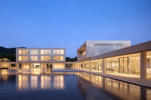 Modern two-story concrete villa with floor-to-ceiling glass walls, illuminated at dusk, featuring a large reflecting pool that mirrors the entire building against a deep blue twilight sky and forested hills.