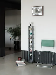 Modern minimalist entryway featuring a green leather folding chair, tall slate-blue shelving unit with ceramics, framed floor plan artwork, and a stack of books with a wooden hippo sculpture on a polished concrete floor.
