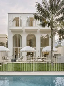 Modern white stucco villa with large arched windows and doors opening to a lush green lawn, outdoor dining area with white umbrellas, and a swimming pool in the foreground under palm trees.