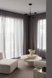 Minimalist living room featuring a large irregular travertine coffee table, modular cream bouclé sofas, layered sheer and opaque grey curtains, and a modern black ceiling light fixture.