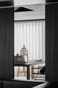 View through a dark wood partition into a bright minimalist home office featuring a wooden easel with calligraphy, stacked books, a simple desk, and a leather chair by the window with sheer white curtains.