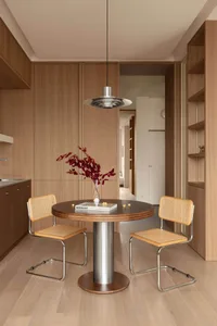 Modern minimalist dining area featuring a round walnut table, two cane chairs with chrome frames, a sculptural metallic pendant lamp, and a vase with red branches against warm wood cabinetry.