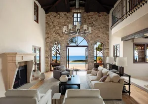 Spacious coastal living room with high vaulted wooden ceiling, textured limestone walls, large beige sectional sofa, modern chandelier, and panoramic ocean view through arched glass doors.