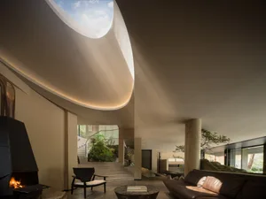 Modern minimalist living room featuring a massive curved skylight, concrete walls, a black fireplace, brown leather sofa, and an indoor courtyard with rocks and trees.