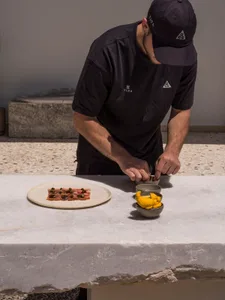 A male chef in black uniform and cap carefully plating food with tweezers on a white marble countertop, with a plate of cured meat and a bowl of yellow garnishes in the foreground.