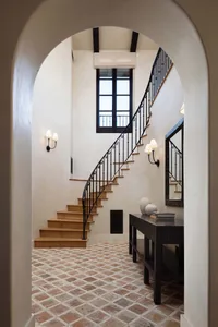 View through a large arched doorway into a spacious entryway featuring a curved wooden staircase with ornate black wrought-iron railing, terracotta tile floor, dark wood console table, and wall sconces.