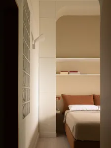 View into a compact modern bedroom featuring a terracotta boucle bed with white linens, built-in arched shelving, and a glass block wall under warm ambient lighting.