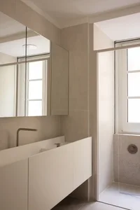Modern minimalist bathroom featuring a floating white vanity, large frameless mirror, micro-cement walls in warm beige, and a walk-in shower with window.